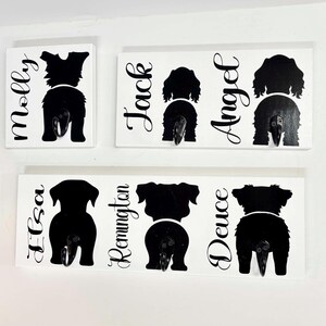 May include: White wooden wall hooks featuring black dog silhouettes with names: Molly, Jack, Angel, Elsa, Remington, and Deuce. Each hook has a metal hook beneath the dog's rear end.
