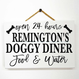 May include: White wooden sign with black text that reads "open 24 hours" with a bone on each side of the text. The sign also reads "Remington's Doggy Diner" and "Food & Water".