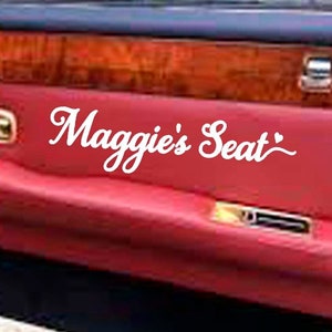 May include: A red leather car door interior with a wood panel and a white vinyl sticker that says "Maggie's Seat".