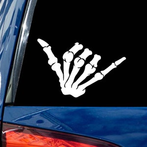 May include: White vinyl decal of a skeleton hand making the shaka sign, a popular Hawaiian gesture of peace, aloha, and good vibes. The decal is perfect for car windows, laptops, or water bottles.