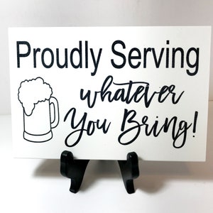 May include: White sign with black text that reads "Proudly Serving whatever you bring!" with a black and white illustration of a beer mug with foam on top.