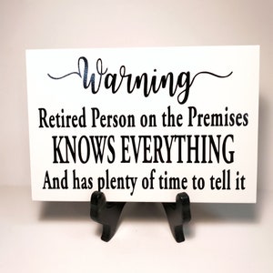 May include: White sign with black text that reads "Warning Retired Person on the Premises KNOWS EVERYTHING And has plenty of time to tell it".
