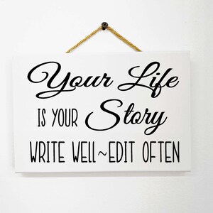 May include: White rectangular wooden sign with black text that reads "Your Life Is Your Story Write Well ~ Edit Often". The sign is suspended by a natural twine rope and a small metal screw.