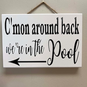 May include: White wooden sign with black text that reads "C'mon around back we're in the Pool" with a black arrow pointing to the right.