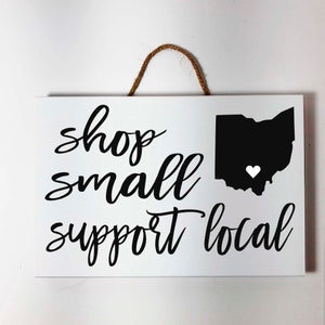 May include: White rectangular wooden sign with the words "shop small support local" in black script. A black silhouette of Ohio with a white heart is on the right. A brown rope is attached for hanging.