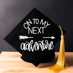 May include: A black graduation cap with the words "ON TO MY NEXT adventure" in white, accompanied by an arrow graphic. It features a gold tassel. The cap rests on a wooden surface, with a dark background.