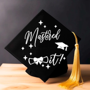 May include: A black graduation cap with white text reading "Mastered it!" and a mortarboard graphic. The cap features a gold tassel. The cap is displayed on a wooden surface.