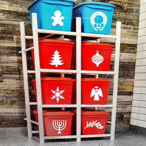 May include: A white shelving unit holds eight plastic storage bins. The bins are red and blue, with white holiday-themed graphics including a gingerbread man, wreath, Christmas tree, ornament, snowflake, angel, menorah, and olive branch.