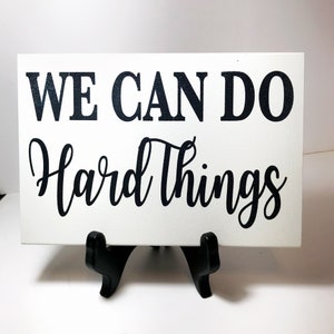 May include: White wooden sign with black text that reads "WE CAN DO Hard Things". The sign is on a black easel.