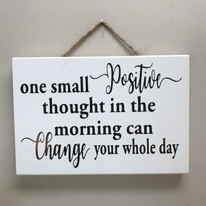 May include: White wooden sign with a black cursive inscription that reads "one small positive thought in the morning can change your whole day".