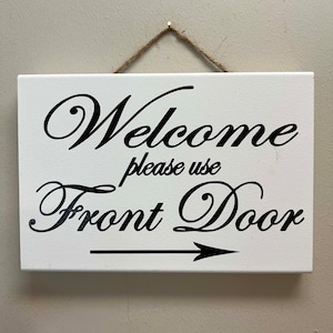 May include: White wooden sign with black text that reads "Welcome please use Front Door" with a black arrow pointing to the right. The sign is hanging from a piece of twine.