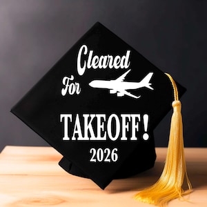 Peut inclure: Coiffe de graduation noire avec l'inscription blanche "Cleared for Takeoff! 2026". Un avion blanc est au-dessus du mot "Takeoff". La coiffe a un gland doré.