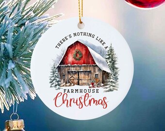 May include: Round Christmas ornament with a farmhouse scene. The ornament features a red barn with a snow-covered roof, evergreen trees, and the text "THERE'S NOTHING LIKE A FARMHOUSE Christmas". It hangs from a gold string.