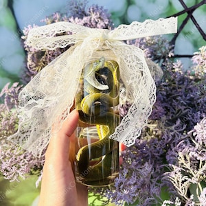 May include: A clear glass jar containing a preserved green snake with its mouth open. The jar is tied with a cream-colored lace ribbon. The background features purple flowers.