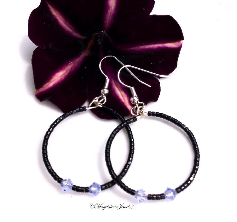 May include: A pair of black beaded hoop earrings with silver hooks and light blue crystal accents. The earrings are displayed against a dark purple flower petal background.