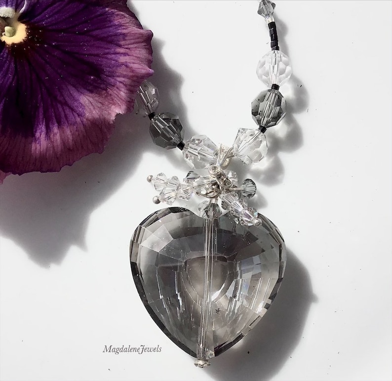 May include: A silver necklace with a large, faceted, clear heart-shaped crystal pendant. The necklace is made with clear and gray crystal beads, and black beads. The pendant is hanging from a silver chain with a silver clasp. The necklace is on a white background.