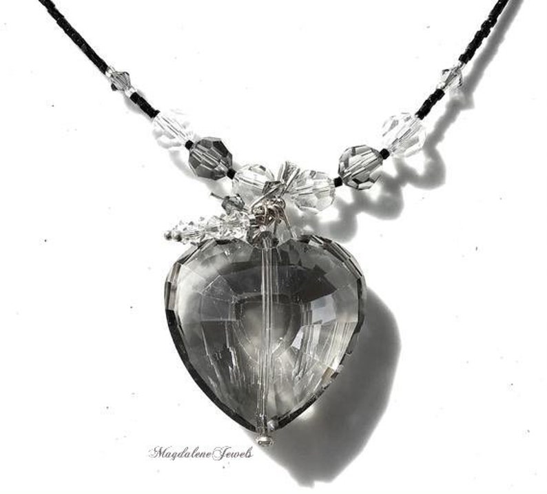 May include: A silver necklace with a large, faceted, clear crystal heart pendant. The necklace is made of black string and has small, clear crystal beads throughout.