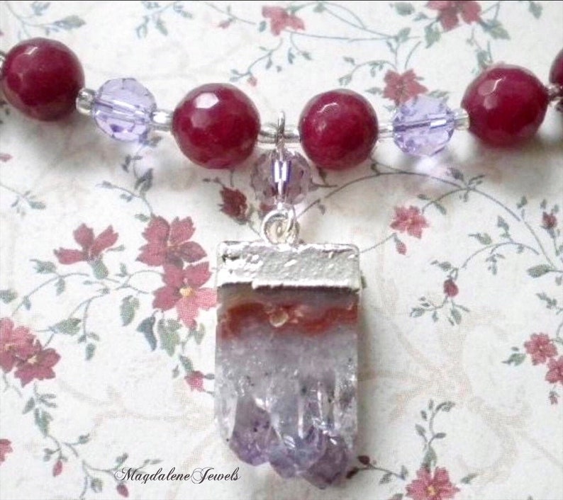 May include: A purple amethyst crystal pendant with a silver setting. The pendant is attached to a necklace made of red beads and purple glass beads.