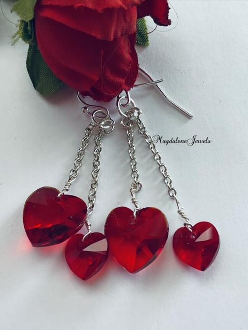 May include: Silver dangle earrings with three red crystal heart charms on each earring. The earrings are displayed against a white background. The text 'Magdalene Jewels' is visible in the image.