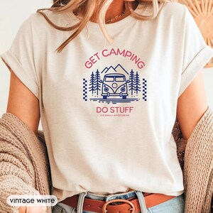Pode incluir: Uma t-shirt branca vintage com uma impress&atilde;o gr&aacute;fica de uma carrinha de campismo, pinheiros e montanhas em azul. O texto diz GET CAMPING DO STUFF e LIVE SMALL + ADVENTURE BIG em rosa e azul. A t-shirt tem mangas curtas e gola redonda.
