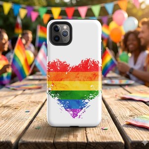 May include: White phone case featuring a distressed rainbow heart design. The heart is composed of horizontal stripes in the colors of the Pride flag: red, orange, yellow, green, blue, and purple. The case is on a wooden table.