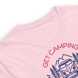 Pode incluir: Uma t-shirt rosa claro com uma impress&atilde;o gr&aacute;fica azul escura de uma carrinha de campismo, pinheiros e montanhas. O texto "GET CAMPING" est&aacute; impresso numa fonte curva rosa escura acima da ilustra&ccedil;&atilde;o, criando um design simples e claro.