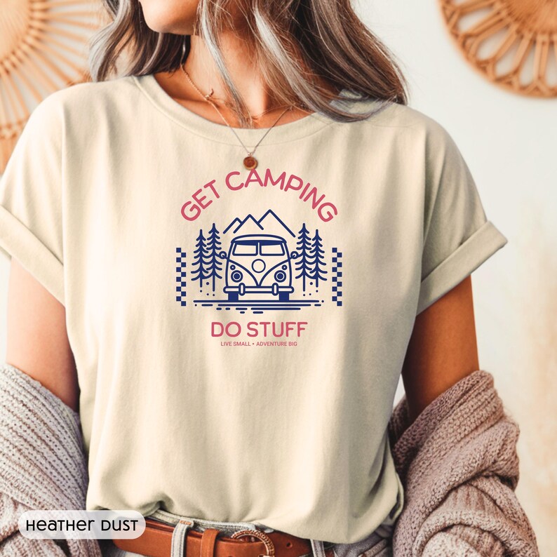 Pode incluir: Uma t-shirt de cor creme com uma impress&atilde;o gr&aacute;fica de uma carrinha azul, pinheiros e montanhas. O texto diz GET CAMPING em rosa, DO STUFF em rosa e LIVE SMALL + ADVENTURE BIG em letras pretas mais pequenas. A t-shirt est&aacute; rotulada como HEATHER DUST.