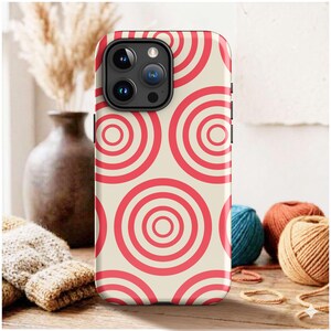 May include: A smartphone case featuring a repeating pattern of concentric circles in bright red against a cream-colored background. The case has a rectangular cutout at the top left for the camera lenses and flash, with a black camera module.