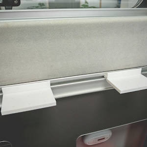 Puede incluir: Dos estantes de plástico blanco montados en una vía de metal plateado. Los estantes están fijados a la vía con tornillos. Los estantes están vacíos.