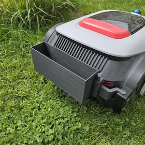 Könnte beinhalten: Ein Roboter-Rasenmäher mit grau-schwarzem Gehäuse, rotem Akzent und einem schwarzen rechteckigen Anbau. Der Mäher steht auf einer grünen Wiese.