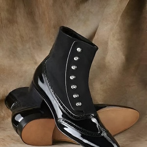 Puede incluir: Botas de tobillo negras de charol con un detalle de botones en el eje. Las botas tienen una punta puntiaguda y un tacón bajo.