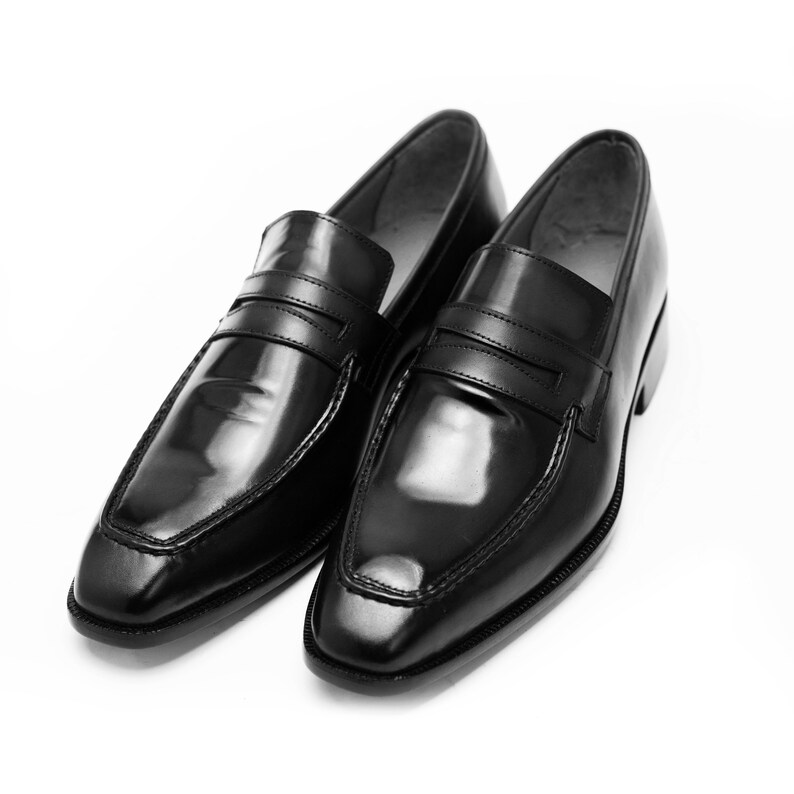 Puede incluir: Un par de mocasines de cuero negro con acabado pulido. Los mocasines tienen una sola correa sobre el empeine y una punta puntiaguda.