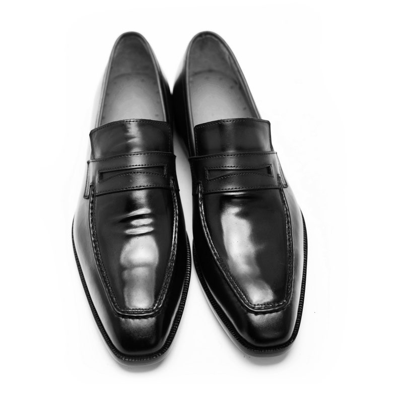 Puede incluir: Un par de mocasines de cuero negro con un detalle de tira de penique. Los zapatos tienen un acabado pulido y una punta puntiaguda.