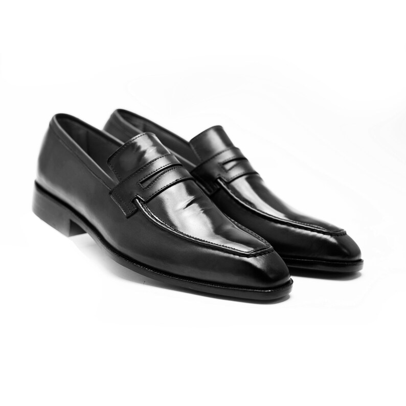 Puede incluir: Un par de mocasines de cuero negro con una sola correa sobre el empeine. Los mocasines tienen un acabado pulido y una punta puntiaguda.