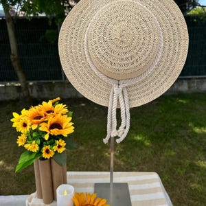 May include: A beige straw sun hat with a wide brim and rope detailing. The hat is displayed on a metal stand. A bouquet of sunflowers and a single sunflower are on a table with a striped beige and white tablecloth.