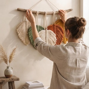 Può includere: Una decorazione da parete con cinque piume in macramè nei colori crema, verde salvia e arancione ruggine, appese a un ramo di legno naturale. Le piume sono realizzate in corda di cotone con nodi dettagliati e bordi sfrangiati.