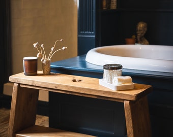 May include: A wooden bench with a shelf, featuring a heart-shaped cutout in the top. The bench is in a bathroom setting with a white bathtub and a dark blue wall.
