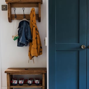 K&ouml;nnte beinhalten: Ein h&ouml;lzerner Garderobenst&auml;nder mit Haken und einem Regal, verziert mit einer sternf&ouml;rmigen Dekoration. Eine blaue Jacke, eine gelbe Jacke mit schwarzem Katzenmuster und eine gr&uuml;ne Spielzeugkamera h&auml;ngen an den Haken. Ein h&ouml;lzerner Schuhst&auml;nder mit zwei Regalen h&auml;lt Schuhe und Hausschuhe. Der Garderobenst&auml;nder steht an einer wei&szlig;en Wand mit einer blauen T&uuml;r rechts.