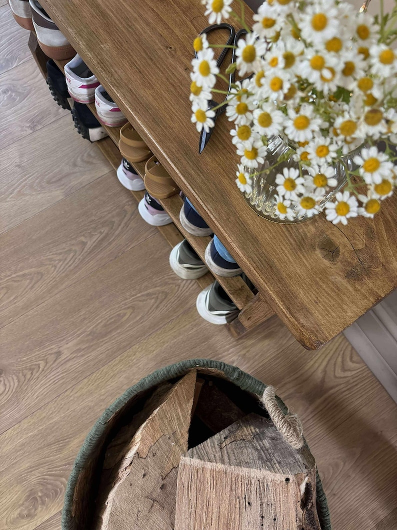May include: A wooden shoe rack with multiple pairs of shoes on the shelves. A vase of white and yellow daisies sits on the top shelf. A basket filled with firewood is in the foreground.