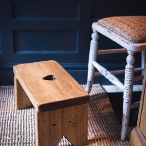 May include: A wooden stool with a heart-shaped cutout in the top. The stool is sitting on a woven rug in front of a white bathtub and a white stool with a floral patterned cushion.