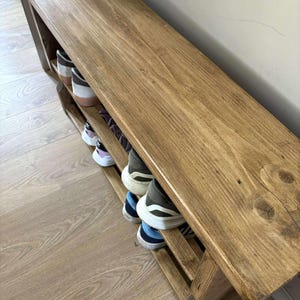 May include: A wooden shoe rack with a bench top. The rack is stained a warm brown color and has two shelves for shoe storage. Several pairs of shoes are visible on the shelves. The bench is approximately 48 inches long.