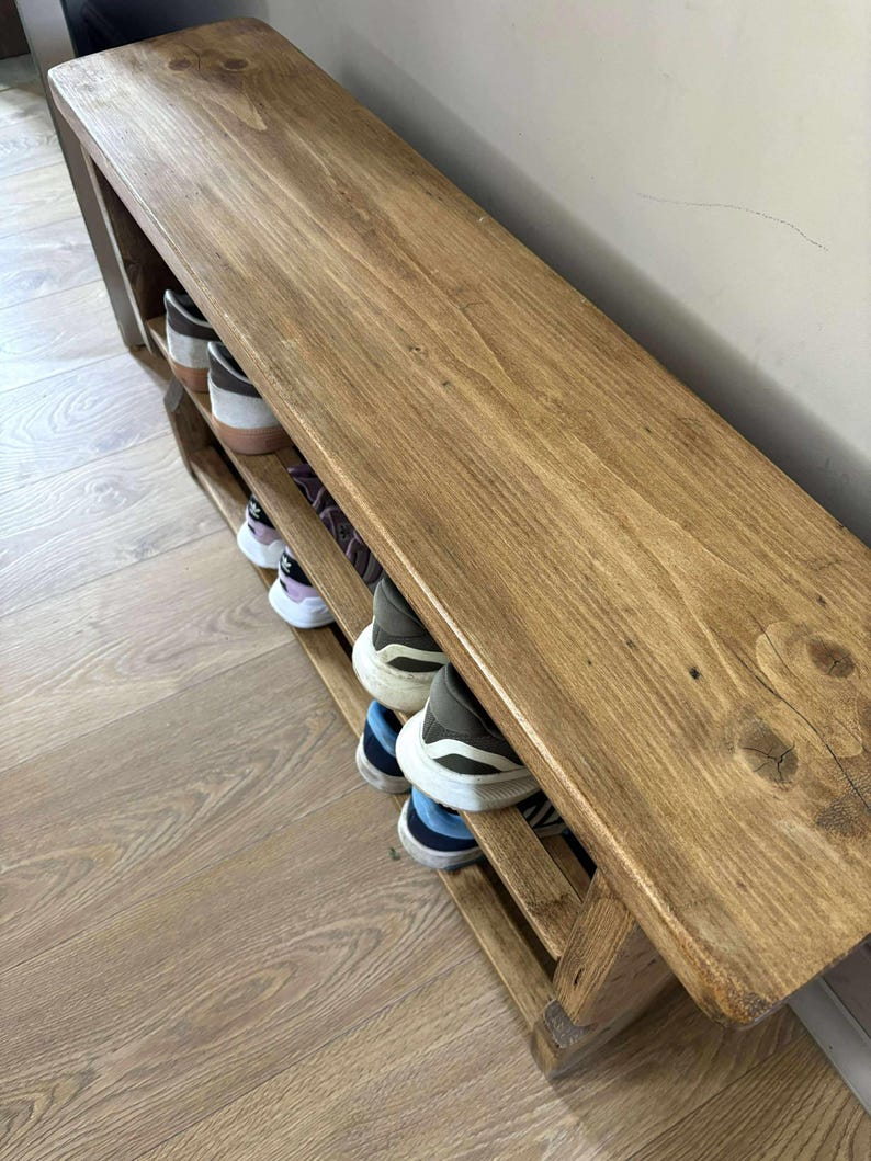 May include: A wooden shoe rack with a bench top. The rack is stained a warm brown color and has two shelves for shoe storage. Several pairs of shoes are visible on the shelves. The bench is approximately 48 inches long.