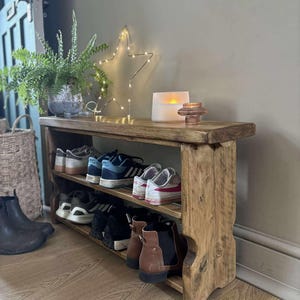 May include: A rustic wooden shoe rack with three shelves, holding various pairs of shoes. The top shelf displays a potted plant, a star-shaped light, and a candle. The rack is made of distressed wood, adding a touch of farmhouse style.
