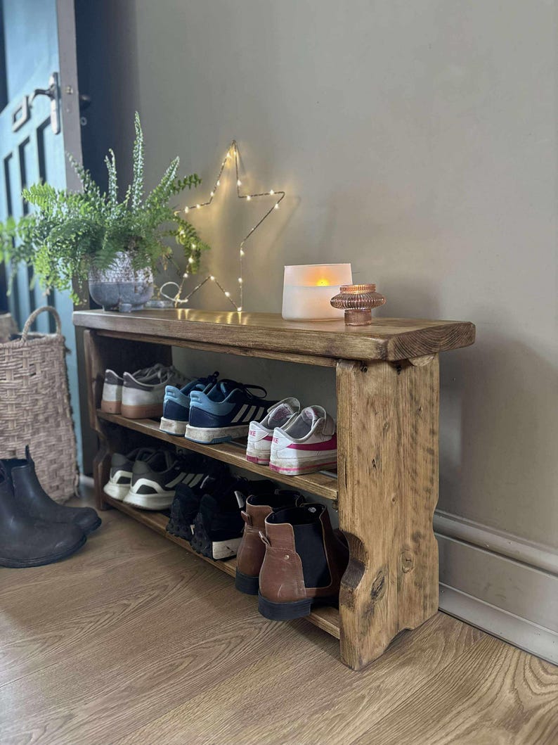 May include: A rustic wooden shoe rack with three shelves, holding various pairs of shoes. The top shelf displays a potted plant, a star-shaped light, and a candle. The rack is made of distressed wood, adding a touch of farmhouse style.