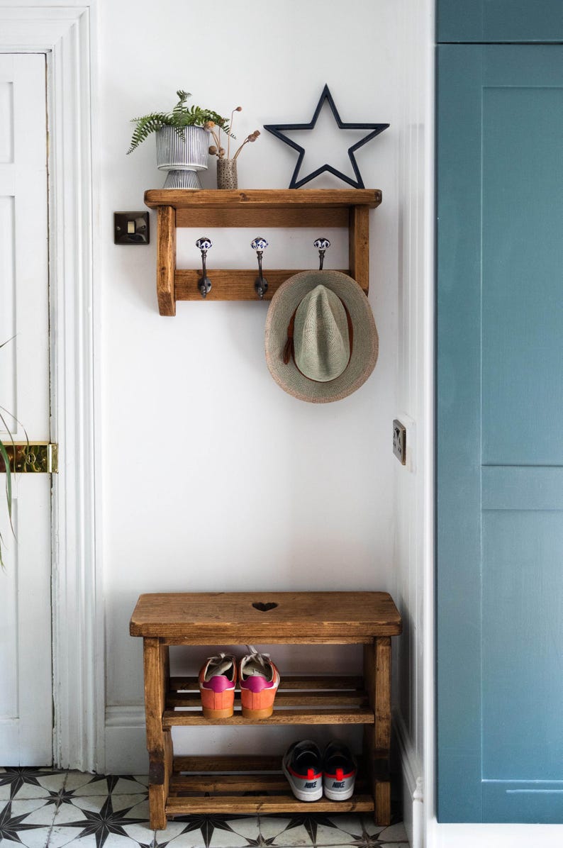 K&ouml;nnte beinhalten: Ein Holz-Kleiderst&auml;nder mit drei Haken und einem Regal. Ein Hut h&auml;ngt an einem der Haken. Ein Holz-Schuhregal mit zwei Regalbrettern befindet sich unter dem Kleiderst&auml;nder. Das Schuhregal hat einen herzf&ouml;rmigen Ausschnitt in der Oberseite.