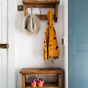K&ouml;nnte beinhalten: Ein Holzregal mit Haken und einer sternf&ouml;rmigen Dekoration dar&uuml;ber. Ein Hut und eine gelbe Jacke h&auml;ngen an den Haken. Eine Holzbank mit zwei Regalen darunter steht in der Ecke. Die Bank hat einen herzf&ouml;rmigen Ausschnitt in der Oberseite. Ein Paar rosa und orangefarbene Turnschuhe und ein Paar schwarze Turnschuhe stehen auf den Regalen.