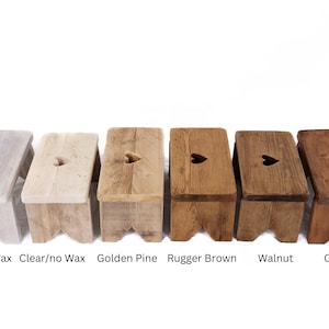 K&ouml;nnte beinhalten: Sechs Holzhocker mit herzf&ouml;rmigen Ausschnitten in der Oberseite. Die Hocker sind in verschiedenen Farben gebeizt: Kalkwachs, klar/ohne Wachs, goldene Kiefer, braunes Leder, Nussbaum und Kirsche.