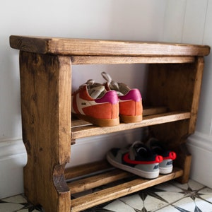 K&ouml;nnte beinhalten: Ein Schuhregal aus Holz mit zwei Ablagen. In der oberen Ablage steht ein Paar orangefarbene und rosafarbene Turnschuhe mit wei&szlig;en Schn&uuml;rsenkeln. In der unteren Ablage steht ein Paar graue und schwarze Turnschuhe mit roten Akzenten.