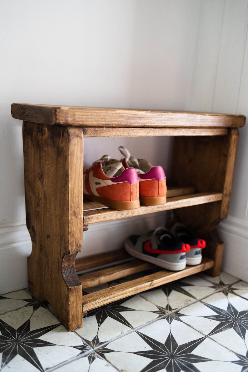 K&ouml;nnte beinhalten: Ein Schuhregal aus Holz mit zwei Ablagen. In der oberen Ablage steht ein Paar orangefarbene und rosafarbene Turnschuhe mit wei&szlig;en Schn&uuml;rsenkeln. In der unteren Ablage steht ein Paar graue und schwarze Turnschuhe mit roten Akzenten.