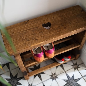 K&ouml;nnte beinhalten: Ein Schuhregal aus Holz mit einem herzf&ouml;rmigen Ausschnitt in der oberen Ablage. Das Regal hat zwei Ablagen und ist in einem dunklen Braunton gebeizt. Zwei Paar Schuhe stehen auf den Ablagen.